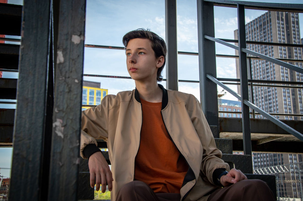 Jeune homme en veste bomber beige assis sur des escaliers métalliques urbains