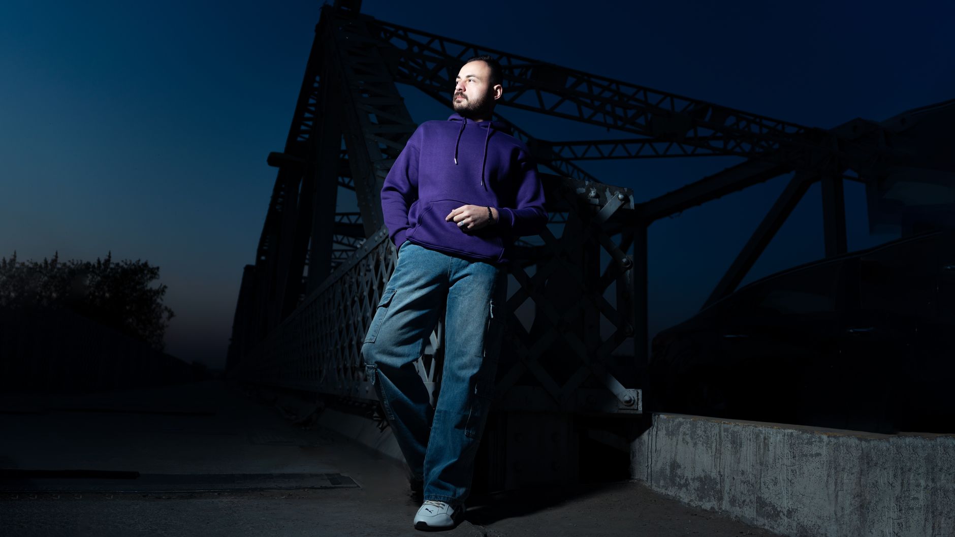 Homme en sweat à capuche violet et pantalon cargo baggy posé sur un pont urbain la nuit