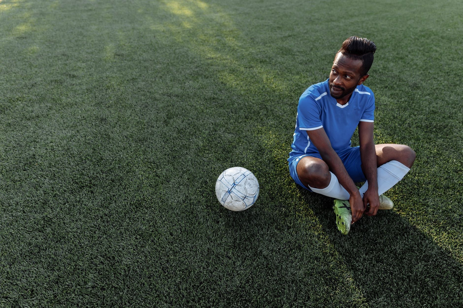 Footballeur assis sur le gazon, tenue bleue, ballon à ses côtés par beau temps