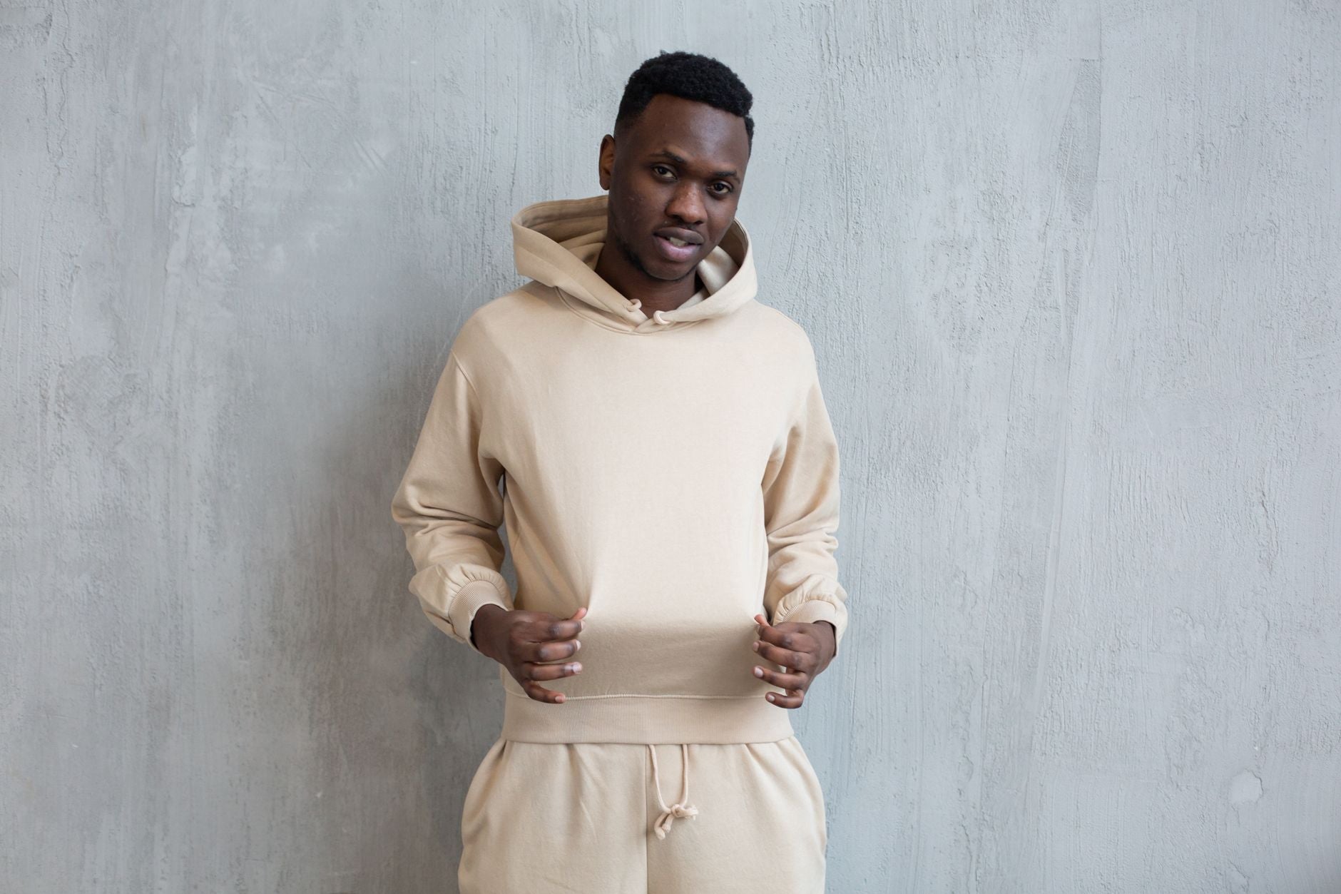 Homme en sweat à capuche beige coordonné, look streetwear urbain épuré contre un mur béton