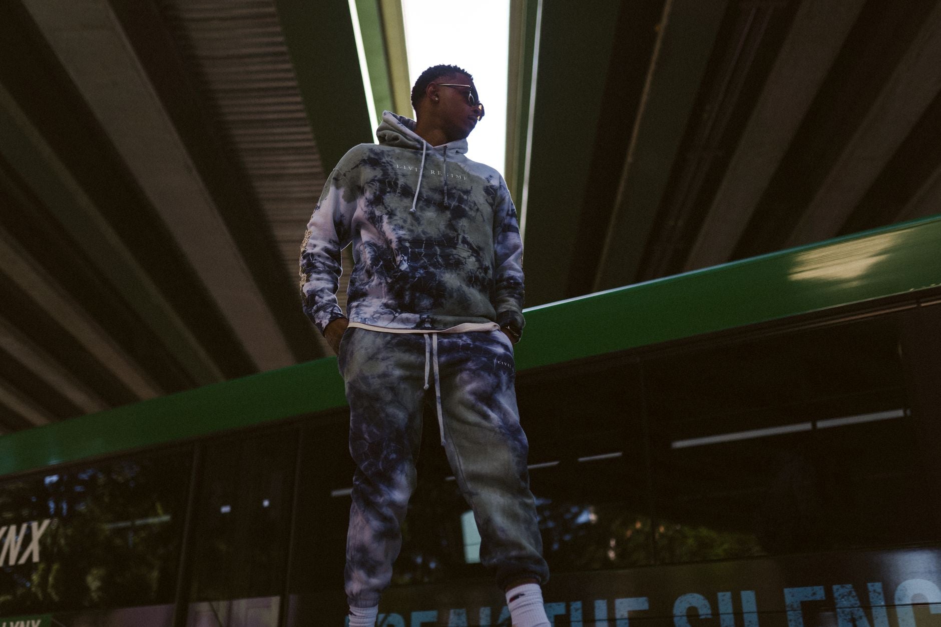 Homme en survêtement streetwear coordonné posant sous un pont urbain