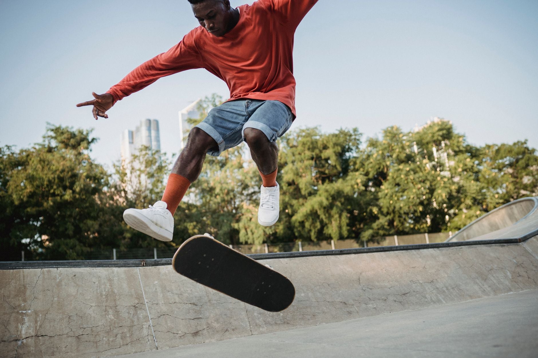 Homme en short en jean et sweat rouge fait un trick de skate en milieu urbain