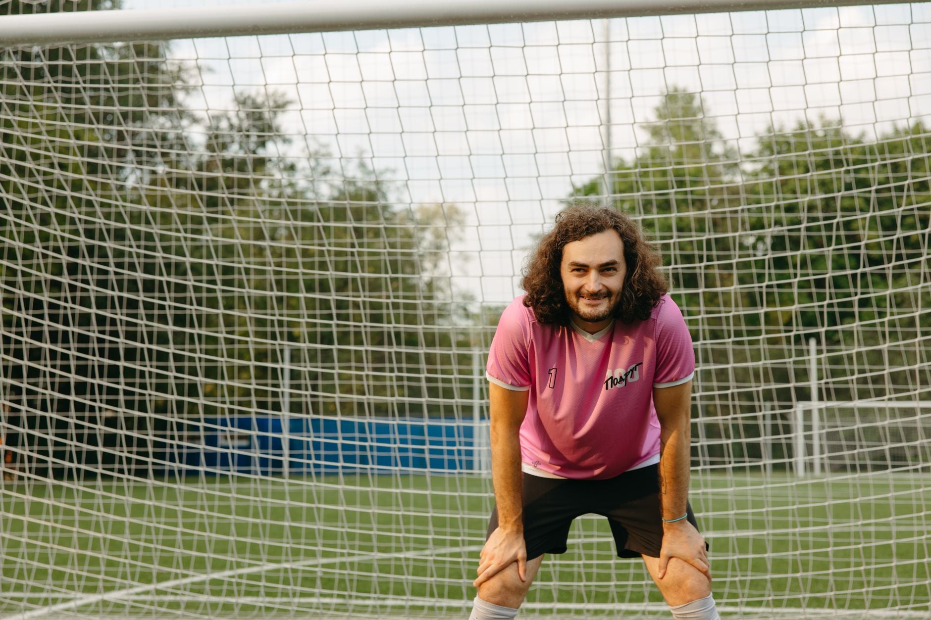 Joueur souriant en maillot de foot rétro rose devant les cages sur terrain extérieur